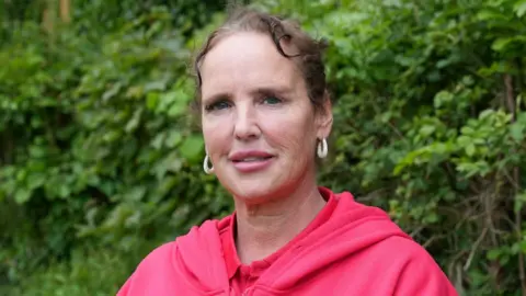 John Fairhall/BBC Sherrie Smith is wearing a pink hoodie in front of a bush. She has brown hair and is wearing glittery earrings.