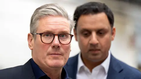 PA Media Sir Keir Starmer in the foreground. He has grey hair and dark-rimmed glasses and is looking straight at the camera. Only his head and shoulders are visible. Anas Sarwar is in the background. He is wearing a blue suit over a white shirt and has dark hair.
