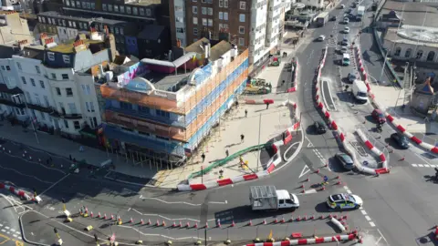 An aerial view of a traffic redevelopment project near a seafront. There are witches hats and barriers placed on the road.