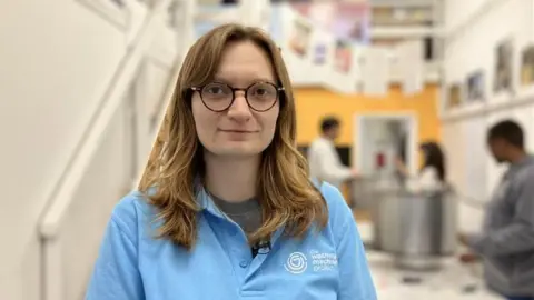 A women is smiling at the camera. She has light brown hair and dark framed glasses. She is wearing a light blue polo top. 