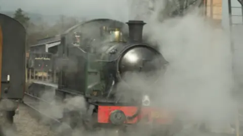 The picture shows a heritage steam locomotive surrounded by thick clouds of steam. The engine is painted in a dark green colour with a red buffer beam at the front, and the number “5526” is visible in gold on the front plate. 