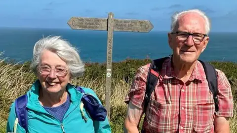 Colin and Doreen Hunt on the coast path. She has a grey-white bob, glasses and is wearing a turquoise windbreaker. He is wearing a checked red shirt and has a navy blue backpack over his shoulders. He has short white hair and black-rimmed glasses