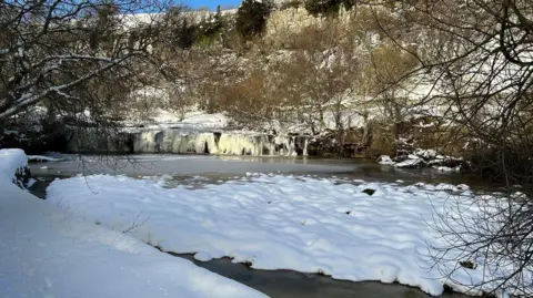 Claire Calvert Wainwath Falls: a small frozen waterfall in the Yorkshire Dales with trees behind it.