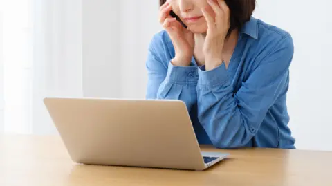 Woman with head in her hands at a laptop on a wooden table. She has shoulder-length dark hair, wearing a blue shirt.