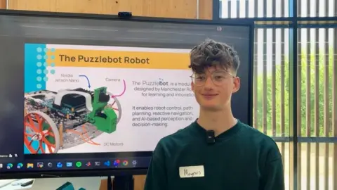 A teenage boy wearing a dark green jumper and glasses stands in front of a large screen. He has short brown hair. Next to the screen is a large window. The screen is displaying an example of how a mini robot works.