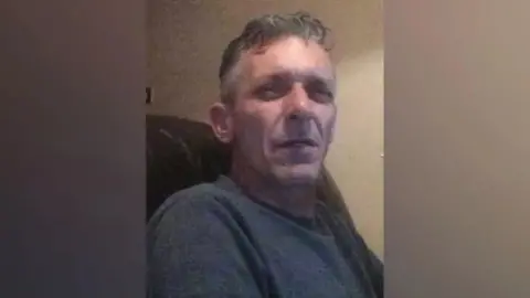South Yorkshire Police A man in his mid-50s with grey hair. He is sitting in an armchair and squinting at the camera.