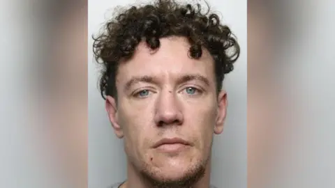 South Yorkshire Police A police mugshot. A man with a long face, blue eyes and tightly curled black hair stands in front of a light grey background.