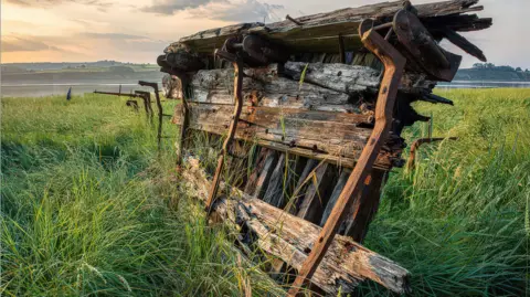 Mark Wardle Part of a wooden boat wreckage sticks out of the grass on a river bank. There are some hills on the other side of the river. It is a sunny evening and the area is glowing in the sun, with a few clouds overhead in the pink-ish sky.