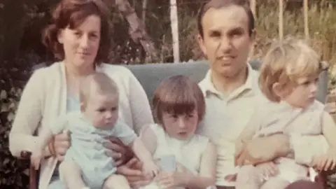 Family photograph A childhood family photograph showing Ali sitting between her parents. Her mother is holding her younger brother, and her father has his arm around her sister.