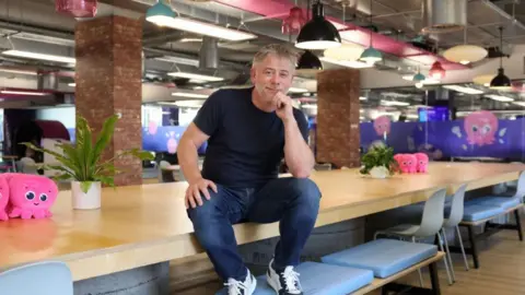Reuters Greg Jackson, founder and CEO of Octopus Energy, poses for a picture on a mess table in the firm's London office. He is wearing a black T-shirt, blue jeans and trainers.