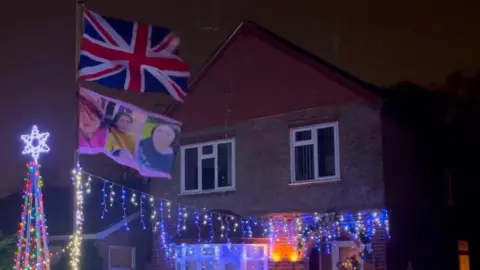 Helen Sadler A Christmas light display outside a house. There are two flags on a flagpole - one is a union jack and the other has three photos of a young girl.