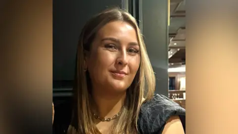 A selfie of a woman with blonde hair smiling. She is wearing a black top and a gold necklace.