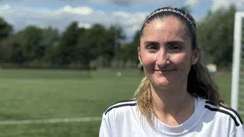A head and shoulders image of a young woman wearing a white and black football shirt. She is standing on a football pitch with a goal behind her. She is smiling and looking into the camera.