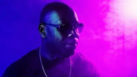 A man standing wearing sunglasses, with half of the background being purple and the other half being pink. He is looking sideways, to the right of the image.