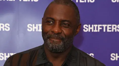 Getty Images Idris Elba wears a black and brown shirt. He is stood in front of a purple background that says "Shifters"