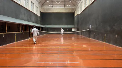 A real tennis court. the floor is large and split halfway by a net. It is surrounded by black walls - some of which are sloped.