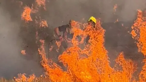 Woburn Community Fire Station Two firefighters tackling a blaze in a field, showing mostly flames and smoke They are wearing helmets but due to the smoke you can hardly see them. 