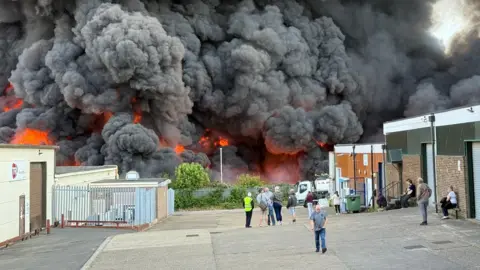 Steve Hubbard/BBC A wall of flames, fire and black smoke are shown in the background while people stand at a distance to survey the scene from a distance in the foreground
