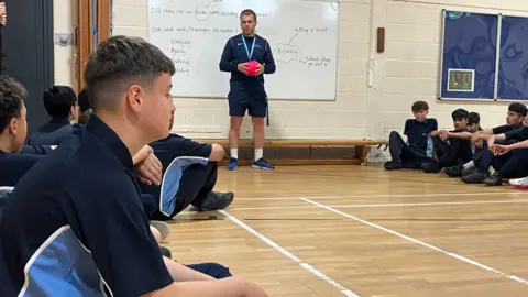 BBC A group of boys sitting in their PE kits on the floor of a basket ball court with a teacher standing beside a white board holding a ball