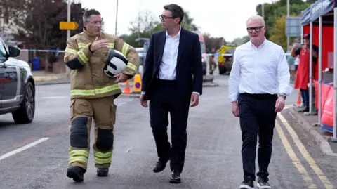 PA Media Andy Burnham, in a dark suit and white shirt, walks down a street alongside chief firefighter Simon Tuhill and the West Midlands mayor Richard Parker
