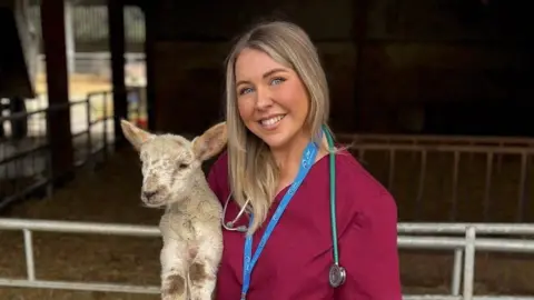 Karis Ramsay holding a lamb in front on an animal pen. Karis is wearing burgundy scrubs, a blue lanyard and ID as well and a stethoscope 