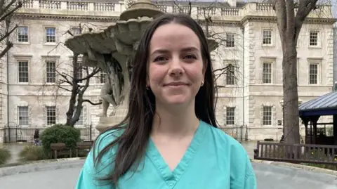PA Media Hollie Page, a young woman with long, brown hair, stands outside a large, old building, wearing green medical scrubs.