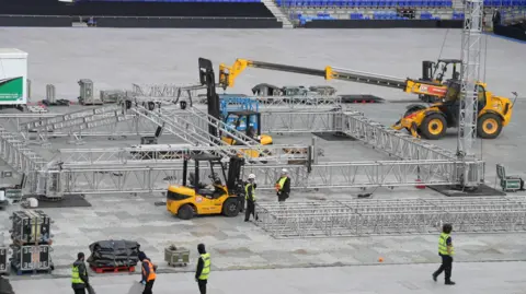 Jamie Niblock/BBC Workers wearing hi-vis jackets walking on the pitch at Portman Road, which is covered in protective plastic boards. There are yellow heavy machinery vehicles putting large metal structures in place where the boxing ring will be.
