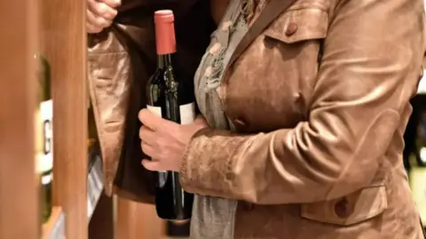 A person hiding a wine bottle under the brown jacket they are wearing