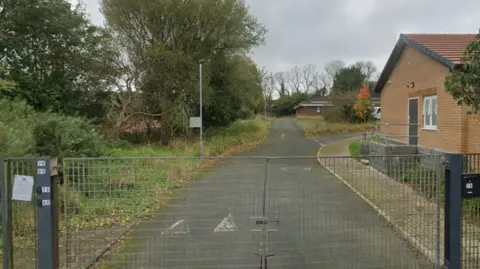 A locked metal gate is closed across a quiet tarmac path leading past a brick building on the right and overgrown greenery and trees on the left, under a cloudy sky.