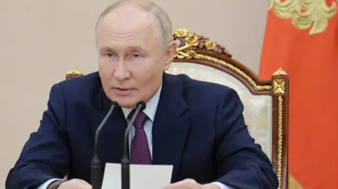 Vladimir Putin sitting on a wooden chair with intricate gold trim, speaking into microphones at the Security Council on 25 September in Moscow. There is a red flag draped behind him and he's holding a small piece of paper.