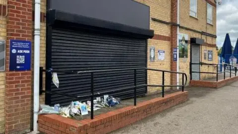 The entrance to the former club shop, which has a small ramp leading to it. The entrance is covered by black shutters. There are a number of flowers left outside of it.