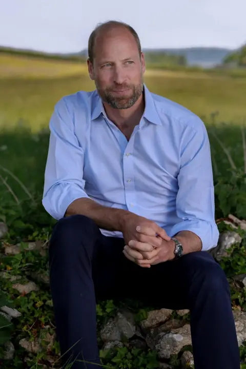Kensington Palace Prince of Wales, wearing a light blue shirt, looks into the distance while sitting on a stone wall with a meadow behind him