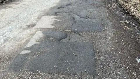 Derek Bennett Ivy House Lane: The left side of the road is light grey, the right side of the road is darker. The right side is pocked with holes and has been patched.
Copyright
Contributed