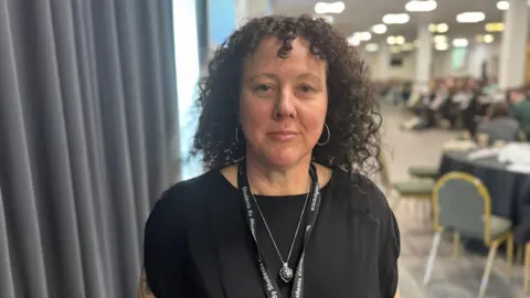 A lady with black curly hair and wearing a black top stands in a conference room with tables of people behind her.