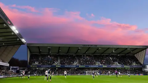Getty Images Millwall in action at the Den in late 2025. The stadium is packed with fans watching the action on the pitch. It is night-time with pink clouds behind one of the stands.