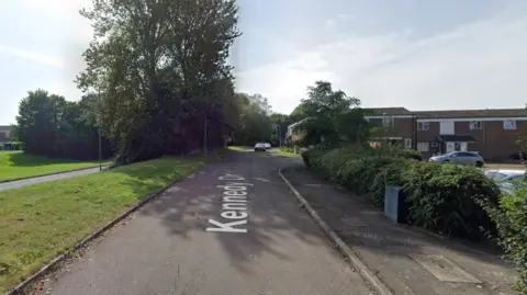 A Google street view image of Kennedy drive - a residential area in Eldene Swindon. There are a number of residential properties and a main road to the left.