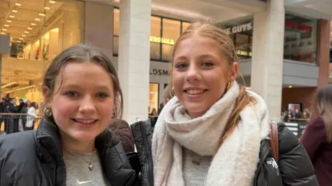 Iris has brown hair tied back and is wearing a grey sweater and black coat. Elsie has ginger hair and is wearing a black coat, white scarf and grey top.