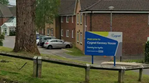 A sign for 'Cygnet Kenney House' in blue and yellow, placed on a small lawn by the entrance to a mental health unit. Cars can be seen parked in a car park while a large tree can be seen on the grass bank.