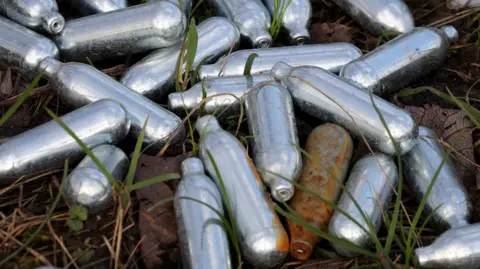 Discarded silver cylindrical nitrous oxide canisters lying on a grassy area, including several that have begun to rust. 