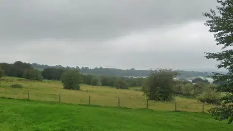 A set of green fields with a fence running through the middle. There are trees in the distance and on the horizon the skyline of a city can be seen.