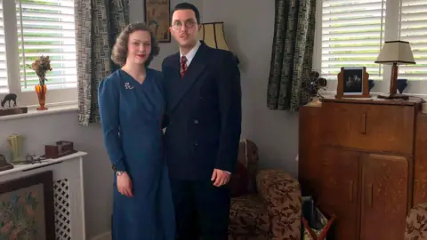 Zoe Dennis/BBC Liberty Avery and Greg Kirby dressed in 1940s clothing in their vintage-styled lounge
