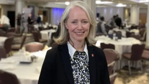 BBC Deputy Kristina Moore smiles at the camera. She is blonde and is wearing a black blazer. There are several tables behind her out of focs like she is at a function.