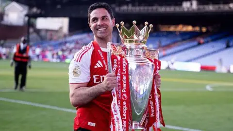 Getty Mikel Arteta smiles as he carries the Premier League trophy across the pitch, with stewards and fans visible in the background.