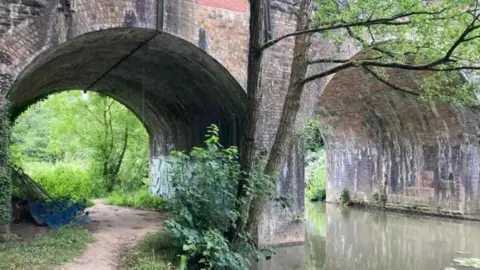 A photo looking under two arches of a bridge over the River Frome and a pathway alongside it which is surrounded by grass and overgrown vegetation. 
