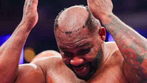 Boxer Jarrell Miller holds his hands to his head as he realises his toupee has fallen off and everyone can see he is bald. His left arm is heavily tattooed.