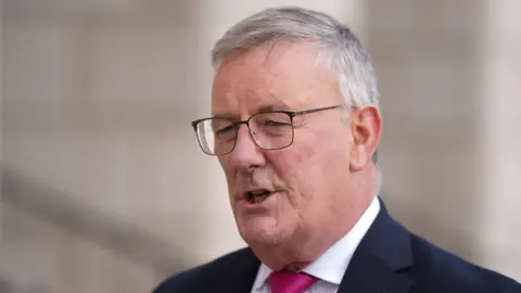 Mike Nesbitt has short grey hair and metal square glasses. He is speaking. He is wearing a white shirt, pink tie and a black blazer.
