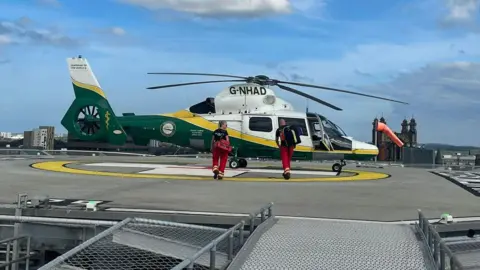 A GNAAS helicopter parked on a helipad with two paramedics walking towards it, carrying medical bags. The skyline of a city is visible behind the helicopter, set against a blue sky with a few white clouds. The helicopter is white, yellow and green. The paramedics are wearing dark t-shirts and red trousers. The helipad is a concrete platform on a rooftop with a yellow and white symbol painted on it where the helicopter is parked.