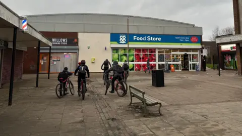 Five young people, seen from behind, ride their bikes through the courtyard of a shopping precinct which is surrounded by shops including a Co-op food store with a blue sign and glass doors and windows. The paved floor of the precinct looks like it needs a makeover. Street furniture includes an old wooden bench and a black bin.