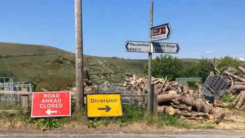 A road sign on the B5470 pointing to Macclesfield in one direction and Whaley Bridge in the other. In front of the signpost, there are temporary signs. One says 'road ahead closed' and is pointing left and the other says 'diversion' and is pointing right.