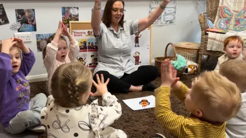A group of children are seated on the carpeted area of a classroom and are taking part in a song. A female teacher is leading the session and is wearing a uniform. 